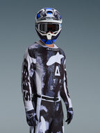 Pilota di motocross con maglia Alpinestars 2026 Racer Lahnd in Iron Camo, caratterizzata da un motivo mimetico nero e grigio e un grande logo Alpinestars bianco sul petto, abbinata a casco e guanti coordinati per il racing fuoristrada.