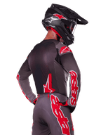 Alpinestars 2025 Supertech Lipan Gear Set, pilota di motocross visto di spalle, maglia e pantaloni grigio antracite con vivace marchio 'astars' rosso sulle maniche e sulla schiena, abbinato a un casco nero