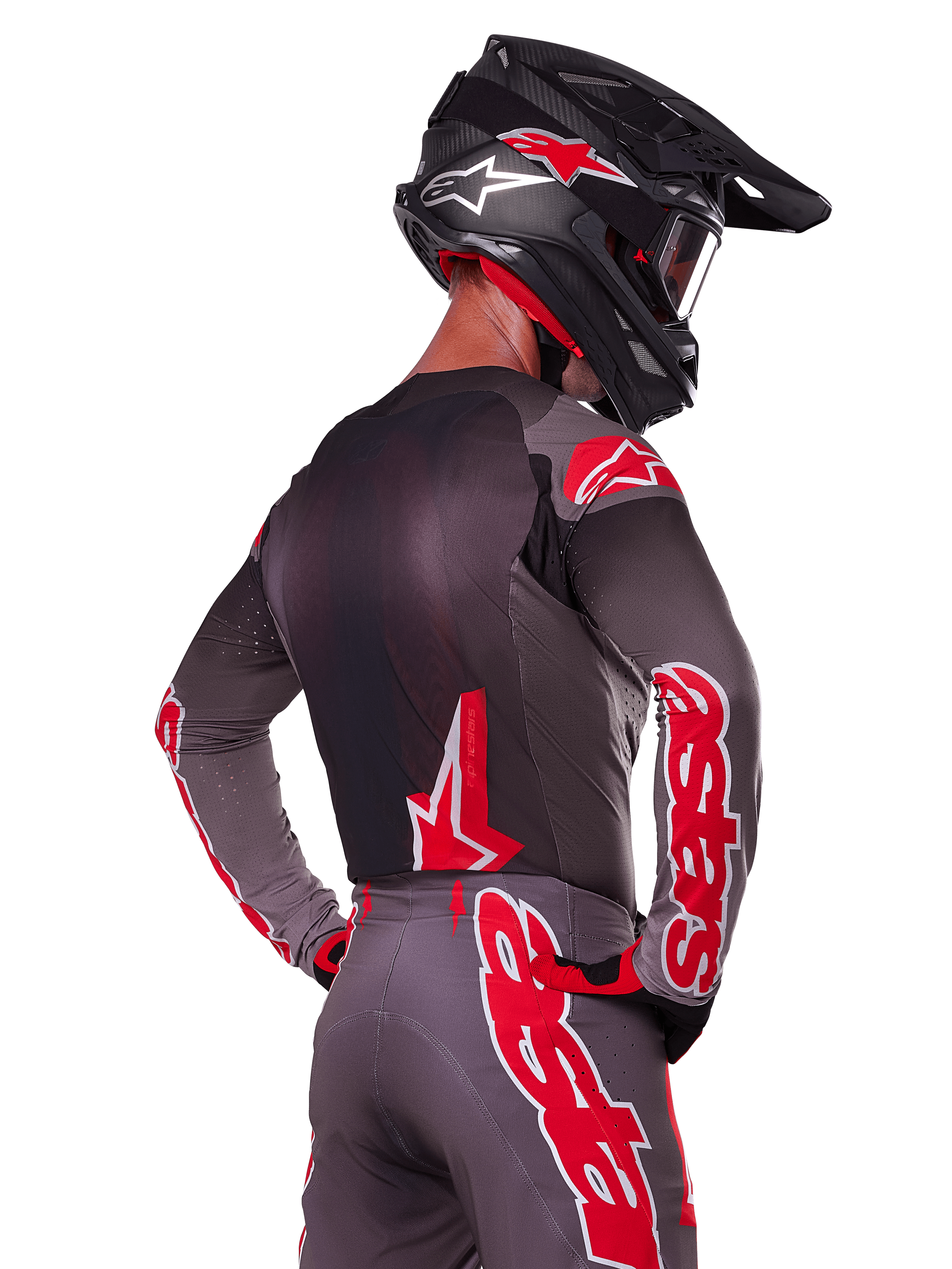 Alpinestars 2025 Supertech Lipan Gear Set, pilota di motocross visto di spalle, maglia e pantaloni grigio antracite con vivace marchio 'astars' rosso sulle maniche e sulla schiena, abbinato a un casco nero