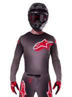 Alpinestars Supertech Lipan Jersey 2025, maglia da motocross, color fumo e rosso, con un grande logo rosso e bianco sul petto, indossata con casco nero e guanti rossi e neri, vestibilità atletica da gara con perforazioni laser