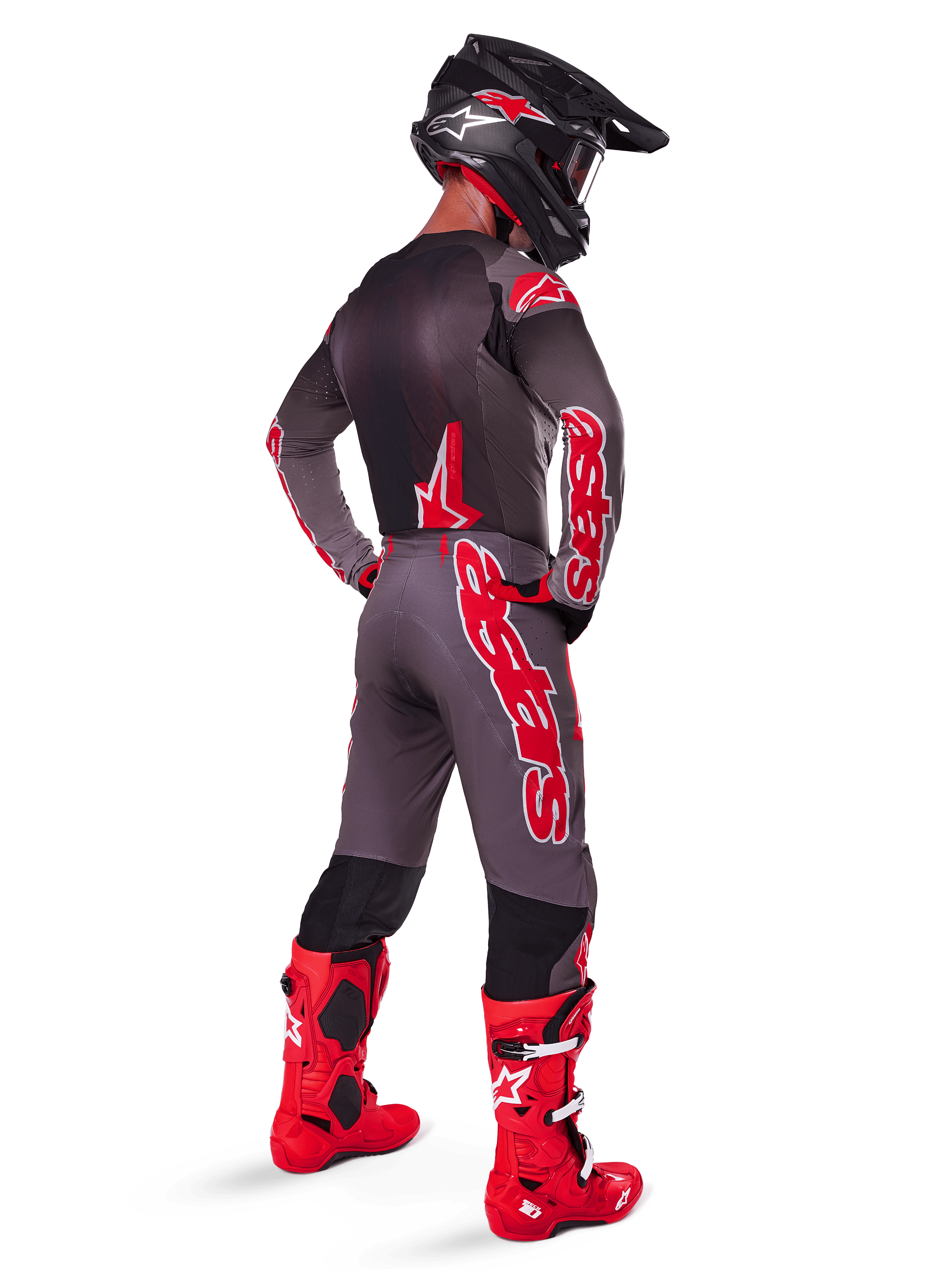 Pilota di motocross con completo Supertech Lipan 2025 in Smoke Bright Red, vista da dietro, maglia grigia e pantaloni coordinati con logo Alpinestars rosso, stivali da gara rosso acceso e casco nero con loghi rossi, abbigliamento professionale completo da fuoristrada