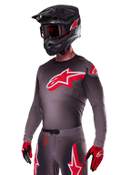 Set Alpinestars 2025 Supertech Lipan, pilota di motocross che indossa maglia e pantaloni grigio scuro con accenti rossi e bianchi, con un grande logo Alpinestars rosso sul petto, perforazioni al laser per la ventilazione, casco nero coordinato, maschera rossa e guanti rossi e neri.
