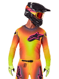 Pilota di motocross con maglia Supertech Lipan 2025, vivace sfumatura gialla, arancione e rosa, grande logo a stella Alpinestars nero con profilo rosa sul petto, perforazioni laser per la ventilazione, vestibilità atletica da gara, abbinata a pantaloni coordinati, guanti gialli e casco nero con maschera.