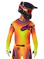 Pilota di motocross con maglia Supertech Lipan 2025, vivace sfumatura gialla, arancione e rosa, grande logo a stella Alpinestars nero con profilo rosa sul petto, perforazioni laser per la ventilazione, vestibilità atletica da gara, abbinata a pantaloni coordinati, guanti gialli e casco nero con maschera.