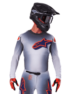 Alpinestars 2025 Supertech Lipan Jersey, maglia da motocross, grigio con accenti arancioni e blu navy, maglia tecnica a maniche lunghe con dettagli di ventilazione traforati, indossata con casco nero e guanti rossi, design racing elegante