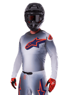 Pilota di motocross con Set Gear Alpinestars 2025 Supertech Lipan, maglia e pantaloni grigi con accenti blu navy e arancioni, casco nero opaco con occhiali e guanti rossi e neri, abbigliamento tecnico per corse fuoristrada