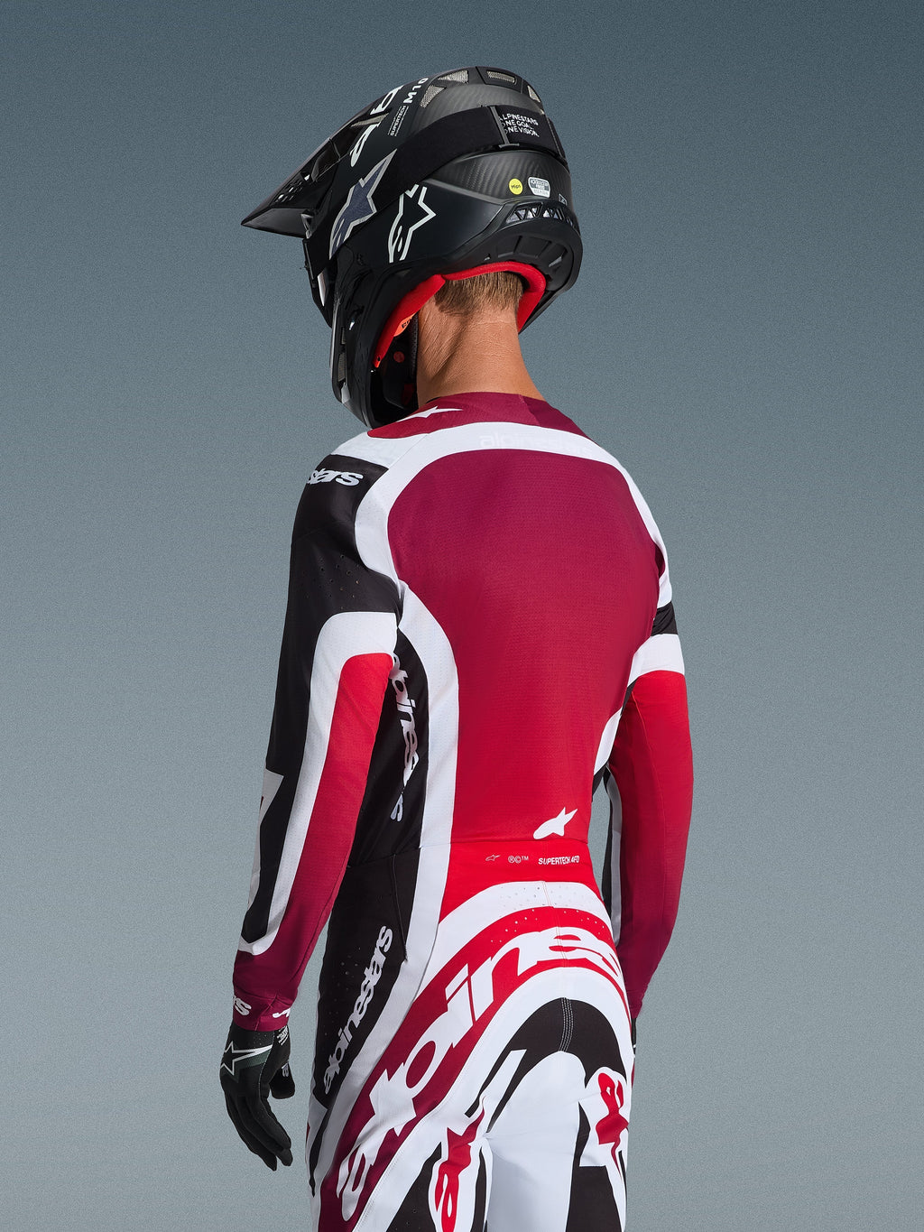 Completo Alpinestars 2026 Supertech AFD Vista, vista posteriore di un pilota di motocross in abbigliamento da gara nero, rosso e bianco, maglia con pannello posteriore rosso scuro e vestibilità atletica, pantaloni coordinati con vistoso logo Alpinestars, casco e guanti neri, tenuta professionale da motocross