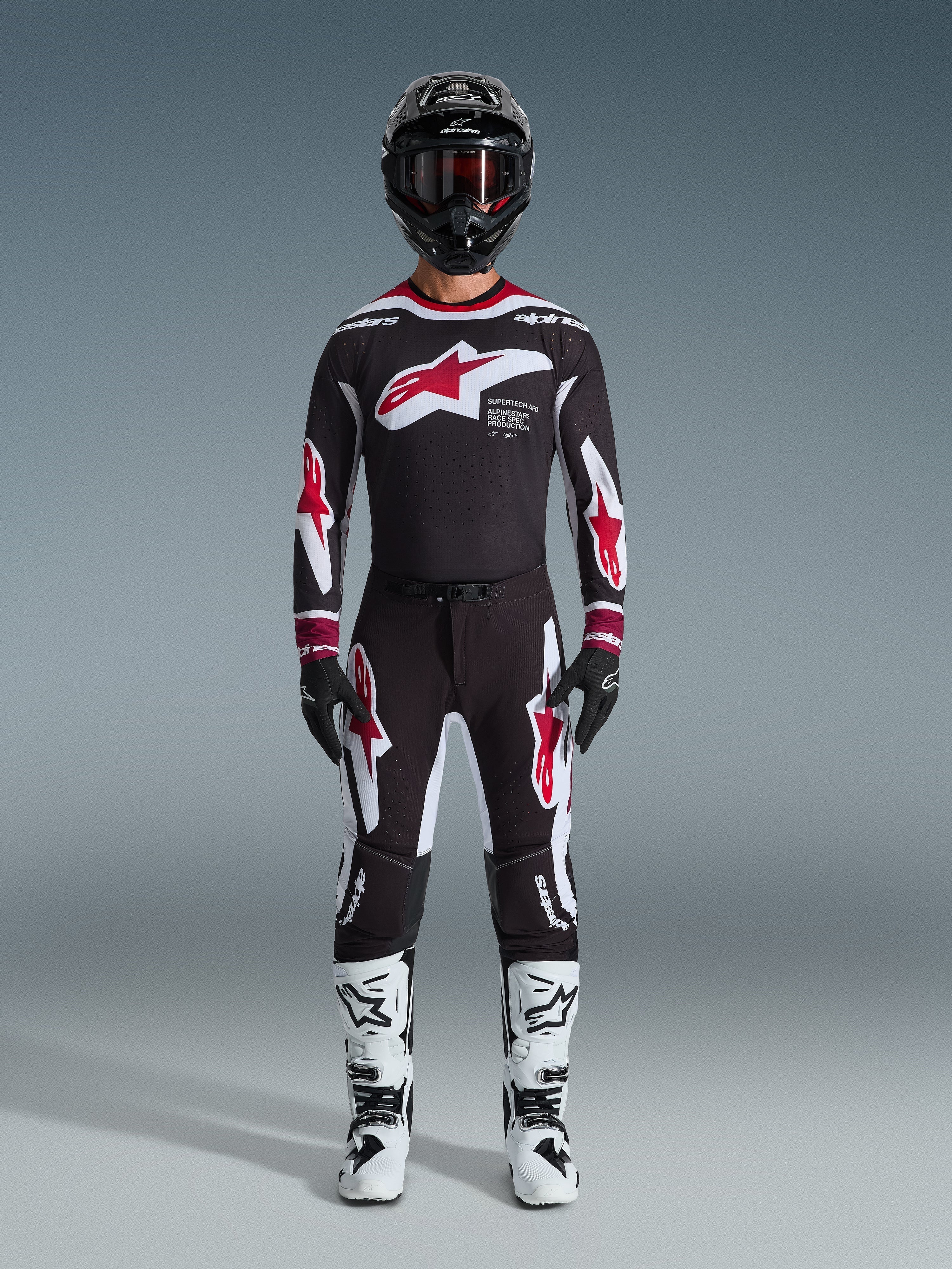 Pilota di motocross con maglia Alpinestars Supertech AFD Vista 2026, completo da gara nero, rosso e bianco, pantaloni neri coordinati con loghi a stella rossi, stivali bianchi e neri, casco nero con occhiali, kit professionale da gara off-road.