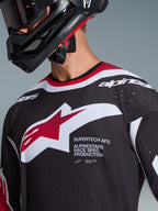 Alpinestars 2026 Supertech AFD Vista Jersey, primo piano di un pilota di motocross che indossa una maglia nera con accenti rossi e bianchi, con un grande logo Alpinestars bianco e rosso, perforazioni di ventilazione tagliate al laser e vestibilità racing professionale, abbinata a un casco nero.
