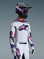 Pilota di motocross con completo Alpinestars Supertech AFD Vista 2026, maglia e pantaloni bianchi con loghi a stella sfumati rosa e viola, casco nero con accenti viola, maschera viola con lenti rosse e guanti bianchi coordinati, abbigliamento professionale per gare fuoristrada