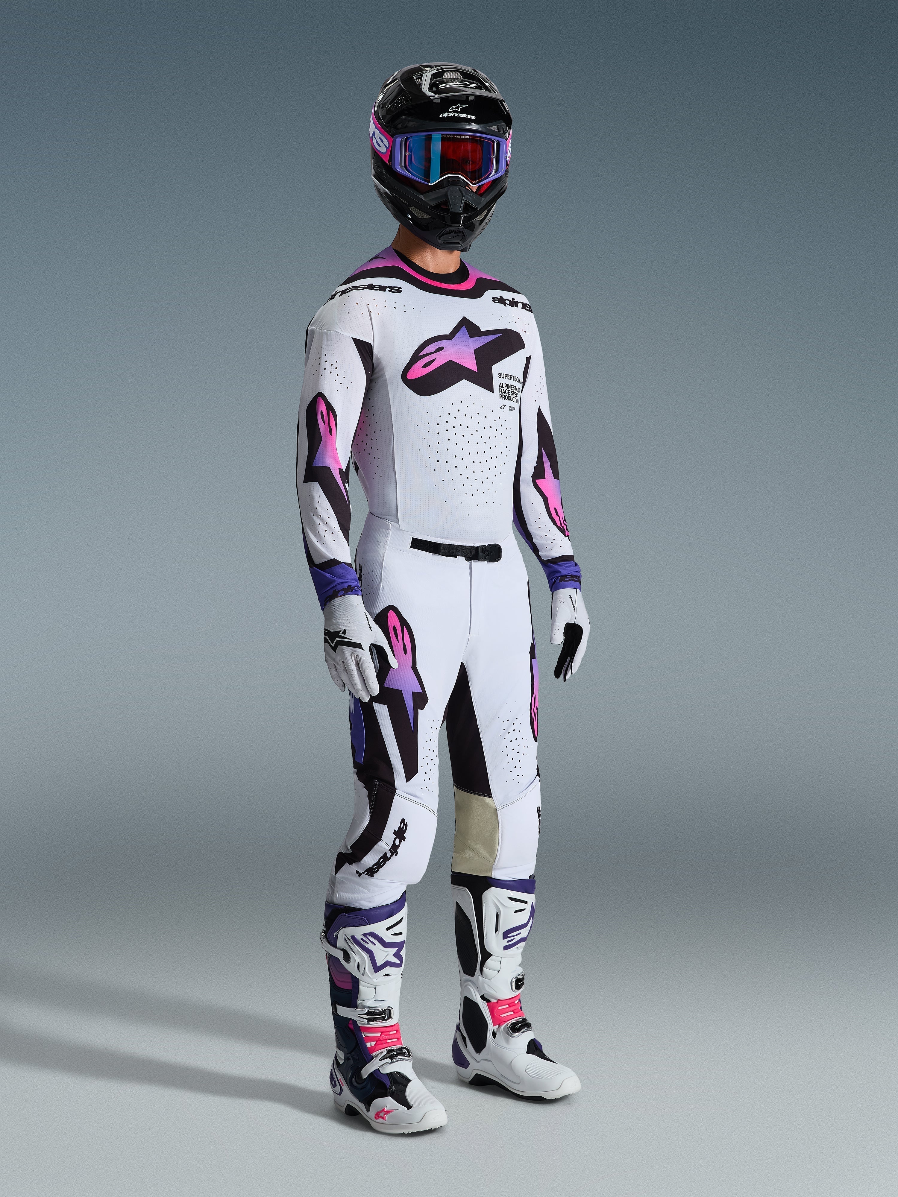 Pilota di motocross che indossa la maglia Alpinestars 2026 Supertech AFD Vista in bianco viola nero, pantaloni da motocross coordinati con loghi sfumati rosa e viola, casco nero con maschera viola, stivali bianchi e neri e guanti bianchi, set completo di abbigliamento professionale da gara off-road
