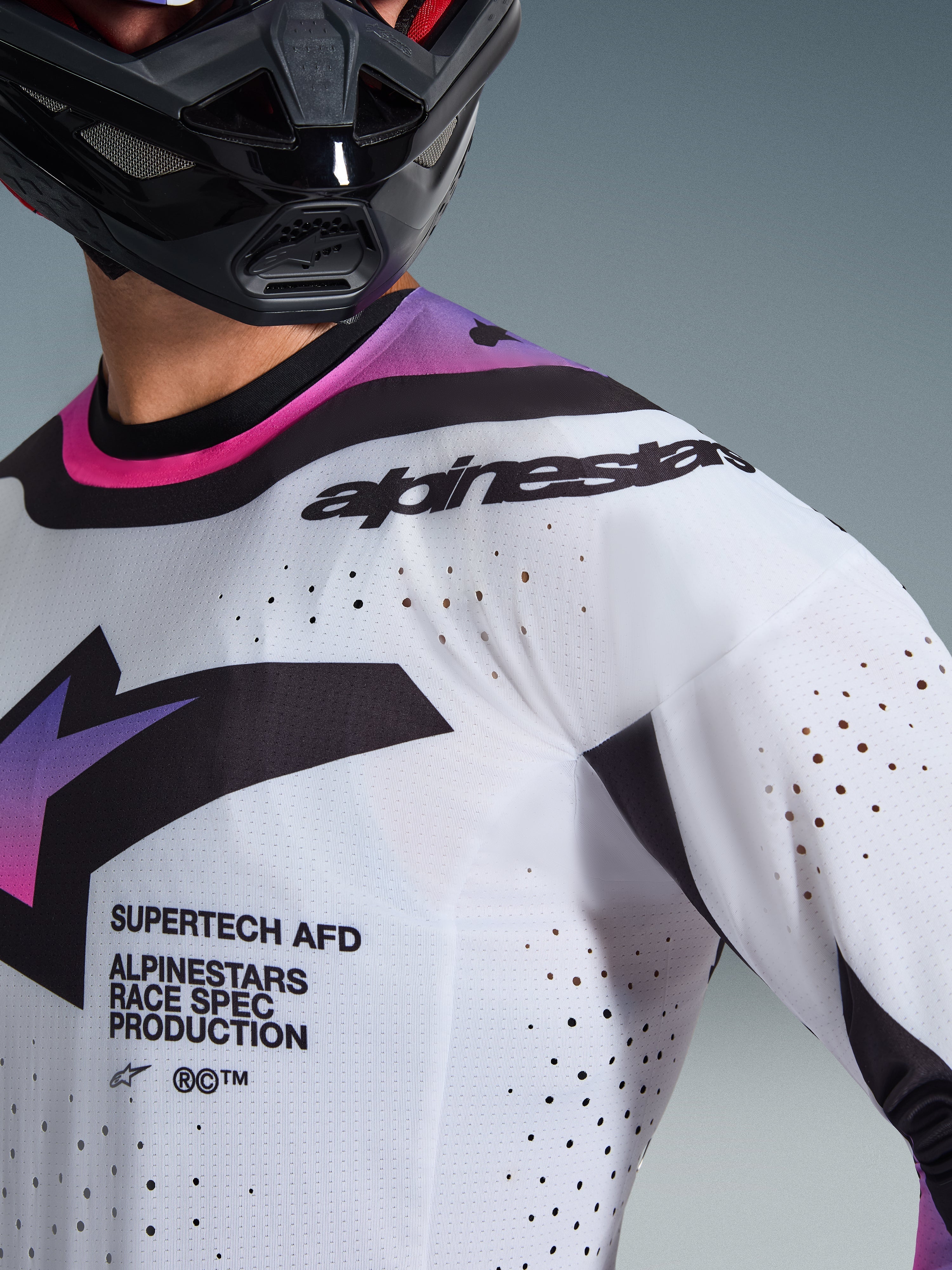 Alpinestars 2026 Supertech AFD Vista Jersey, maglia da motocross, bianco, viola e nero, vista ravvicinata che mostra le perforazioni tagliate al laser e il branding race spec, indossata con un casco nero, abbigliamento professionale da gara fuoristrada
