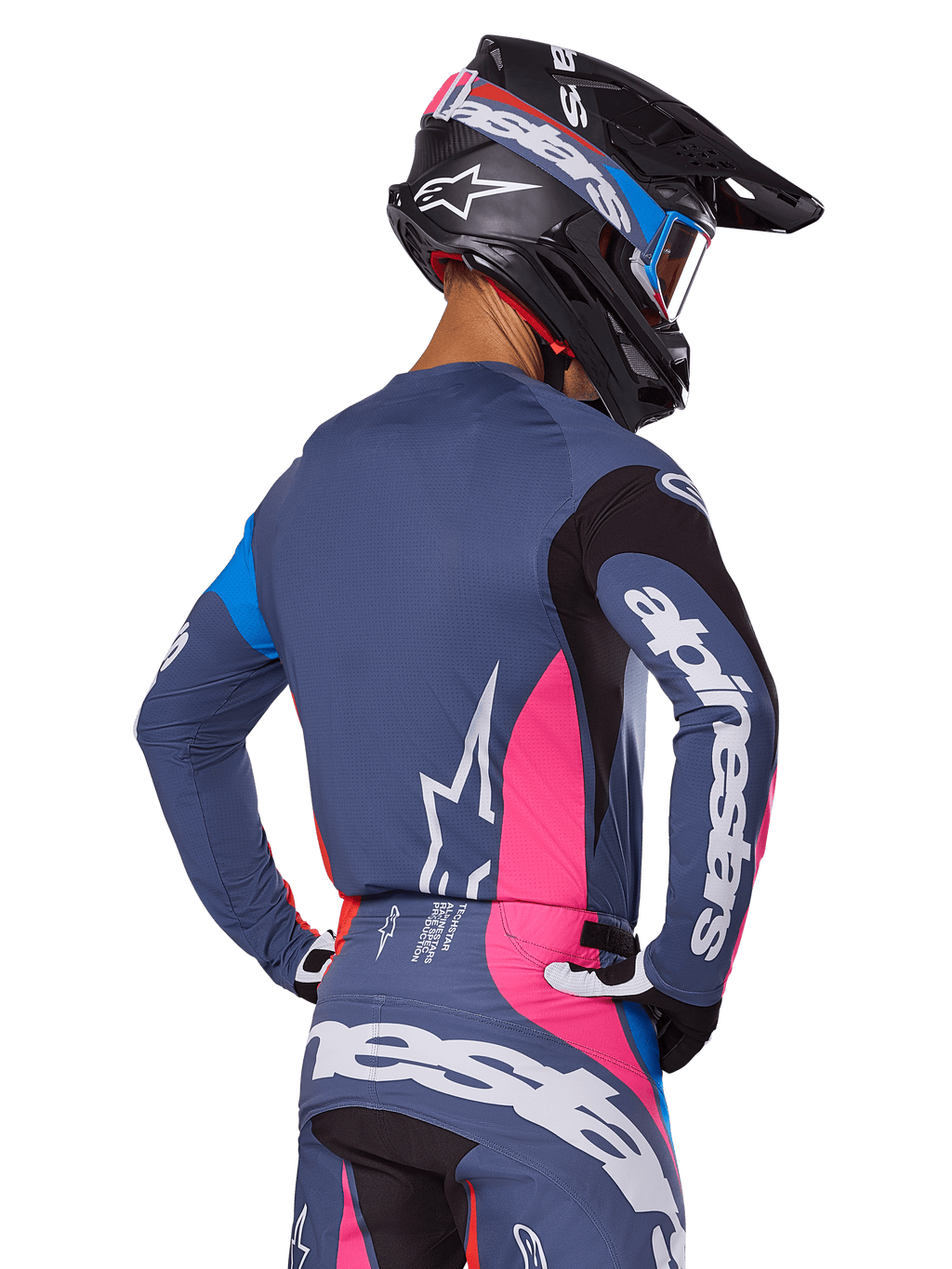 Pilota di motocross visto da dietro con set di equipaggiamento Alpinestars Techstar Dreem 2025 in grigio, maglia e pantaloni blu-grigio con pannelli rosa e neri, loghi Alpinestars bianchi su maniche e schiena e casco nero coordinato con grafiche blu e rosa.