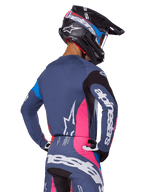 Pilota di motocross visto da dietro con set di equipaggiamento Alpinestars Techstar Dreem 2025 in grigio, maglia e pantaloni blu-grigio con pannelli rosa e neri, loghi Alpinestars bianchi su maniche e schiena e casco nero coordinato con grafiche blu e rosa.
