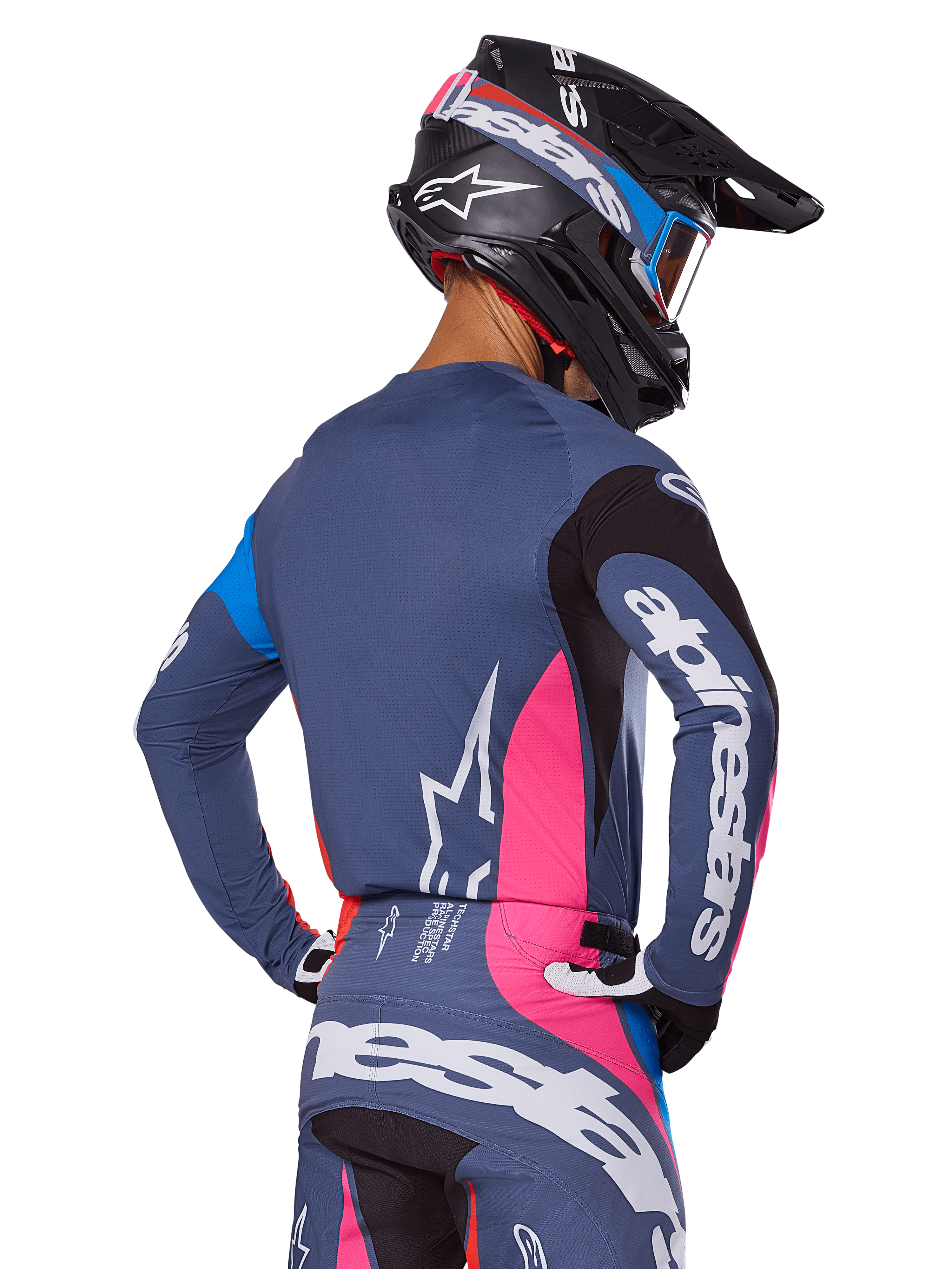 Pilota di motocross visto da dietro con set di equipaggiamento Alpinestars Techstar Dreem 2025 in grigio, maglia e pantaloni blu-grigio con pannelli rosa e neri, loghi Alpinestars bianchi su maniche e schiena e casco nero coordinato con grafiche blu e rosa.