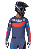 Alpinestars maglia Techstar Dreem 2025, maglia da motocross, grigio con accenti blu, rossi e rosa, con perforazioni laser e grande logo sul petto, indossata con pantaloni coordinati e casco da gara nero