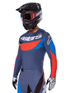 Completo motocross Alpinestars Techstar Dreem 2025, pilota che indossa maglia e pantaloni grigio ardesia con grafiche rosse e blu e logo 'astars' sul petto, abbinato a casco nero, maschera con montatura rossa e guanti da gara bianchi