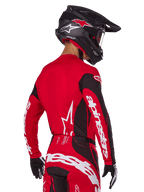 Pilota di motocross visto da dietro con completo Alpinestars Techstar Dreem 2025, maglia rossa e nera con loghi bianchi sulle maniche, pantaloni coordinati rossi e neri con logo bianco sulla seduta, casco in carbonio nero con accenti rossi, guanti bianchi e neri, abbigliamento professionale da gara fuoristrada