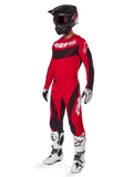 Vista a figura intera di un pilota di motocross con il completo Techstar Dreem 2025 in rosso brillante e nero, con maglia tecnica con branding bianco, pantaloni coordinati in ripstop elasticizzato, casco nero con visiera rossa, guanti bianchi e neri e stivali Tech bianchi, abbigliamento da gara specializzato per competizioni fuoristrada.