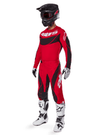 Vista a figura intera di un pilota di motocross con il completo Techstar Dreem 2025 in rosso brillante e nero, con maglia tecnica con branding bianco, pantaloni coordinati in ripstop elasticizzato, casco nero con visiera rossa, guanti bianchi e neri e stivali Tech bianchi, abbigliamento da gara specializzato per competizioni fuoristrada.