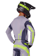 Pilota di motocross visto da dietro che indossa il set Alpinestars Techstar Dreem 2025, maglia e pantaloni grigi con vivace branding giallo-verde neon, casco nero coordinato con grafiche neon, completo professionale da motocross