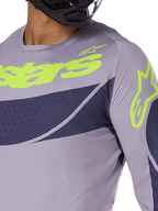 Alpinestars Techstar Dreem Jersey 2025, maglia da motocross, grigio e blu navy con logo verde lime acceso, primo piano del pilota con casco nero, con perforazioni tagliate al laser per la ventilazione