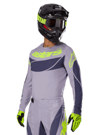 Pilota di motocross che indossa la maglia Alpinestars 2025 Techstar Dreem in grigio e grigio scuro, con logo 'astars' giallo neon sul petto, completata da casco, maschera e guanti coordinati per un look da gara off-road.