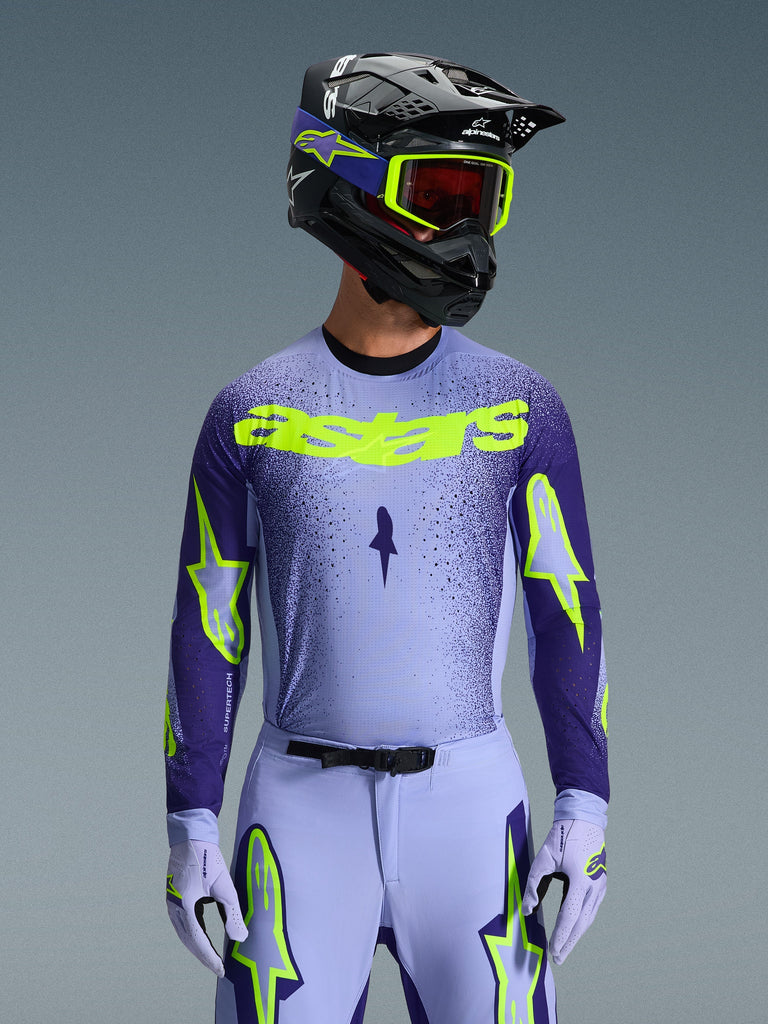 Maglia Alpinestars Supertech Scenz 2026, maglia da motocross, viola e giallo fluo, indossata da un pilota con casco coordinato nero e viola, occhiali giallo neon e pantaloni da corsa viola, con marchio astars giallo neon sul petto e loghi a stella sulle maniche.