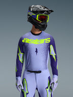 Vista frontale di un pilota di motocross con il Completo Supertech Scenz 2026 di Alpinestars, maglia viola chiaro e blu scuro con logo 'astars' giallo neon, pantaloni e guanti coordinati viola chiaro con accenti neon, casco nero con occhiali giallo neon, scatto in studio su sfondo grigio