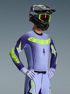 Pilota di motocross con maglia Alpinestars Supertech Scenz 2026, viola e giallo fluo, pantaloni viola abbinati con accenti fluo, casco nero con occhiali gialli e guanti da gara viola, attrezzatura professionale coordinata per corse fuoristrada