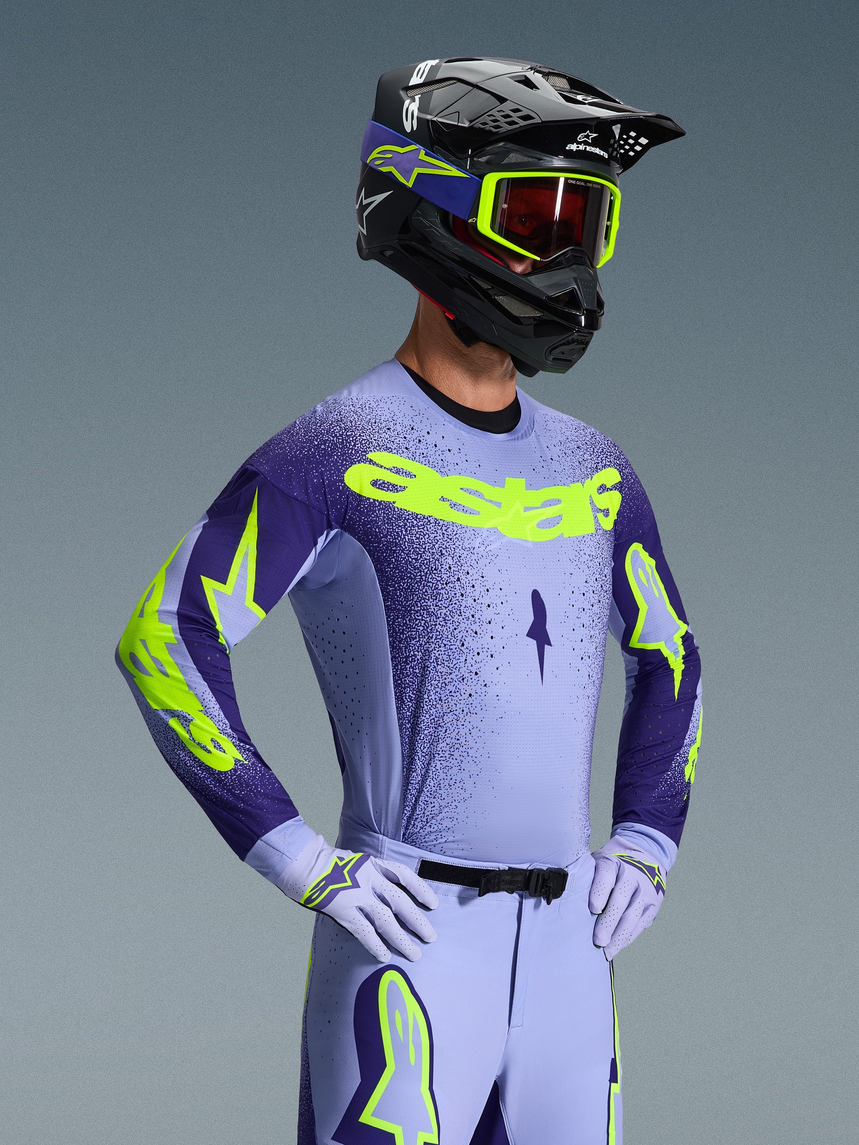 Pilota di motocross con maglia Alpinestars Supertech Scenz 2026, viola e giallo fluo, pantaloni viola abbinati con accenti fluo, casco nero con occhiali gialli e guanti da gara viola, attrezzatura professionale coordinata per corse fuoristrada