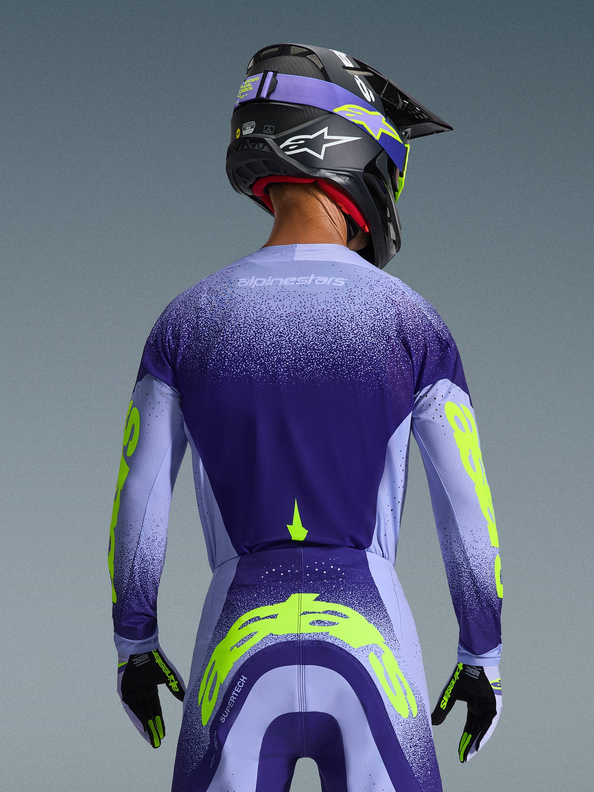 Maglia Alpinestars Supertech Scenz 2026 in viola e azzurro con accenti giallo fluo, vista posteriore del pilota con pantaloni da motocross coordinati, casco nero e guanti, abbigliamento tecnico ad alte prestazioni per gare fuoristrada
