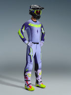 Pilota di motocross con maglia Alpinestars Supertech Scenz 2026 e kit da gara completo, design viola e giallo fluo con base viola chiaro e accenti neon, pantaloni coordinati con loghi a stella, casco nero con maschera giallo neon, stivali viola e giallo neon, abbigliamento professionale da fuoristrada