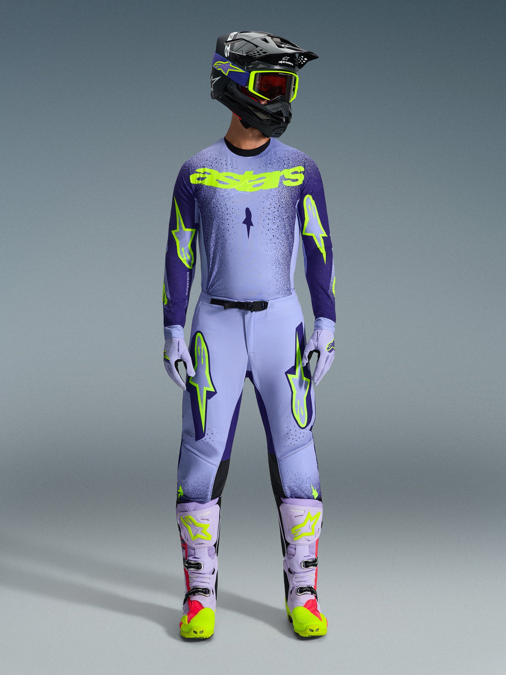 Pilota di motocross con abbigliamento completo Alpinestars Supertech Scenz 2026, maglia viola e giallo fluo con pantaloni abbinati, casco nero con occhiali gialli, stivali viola chiaro e neon, completo professionale per corse off-road