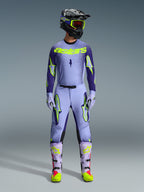Pilota di motocross con abbigliamento completo Alpinestars Supertech Scenz 2026, maglia viola e giallo fluo con pantaloni abbinati, casco nero con occhiali gialli, stivali viola chiaro e neon, completo professionale per corse off-road