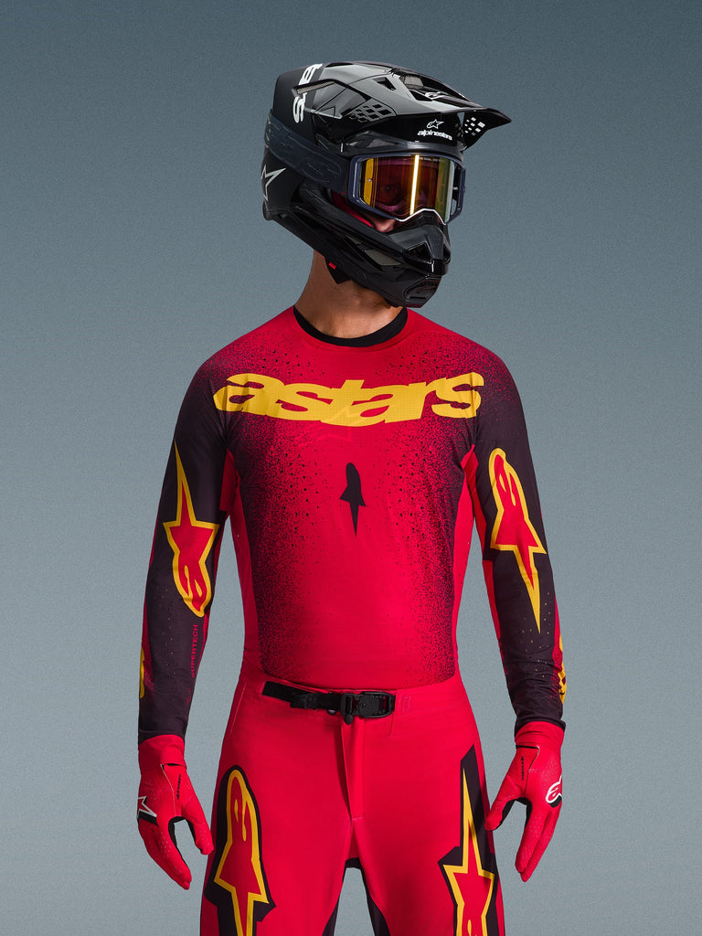 Alpinestars 2026 Supertech Scenz Jersey, maglia da motocross, rosso, nero e giallo, indossata da un pilota con casco nero coordinato e guanti rossi, con un vistoso logo 'astars' giallo sul petto e un motivo punteggiato nero sfumato sulle spalle.