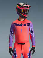 Pilota di motocross con completo Alpinestars Supertech Scenz 2026, maglia rosa-arancio vivace con maniche viola e branding 'astars', pantaloni coordinati con dettagli a stella viola, casco multicolore con maschera, guanti neri, abbigliamento professionale per fuoristrada