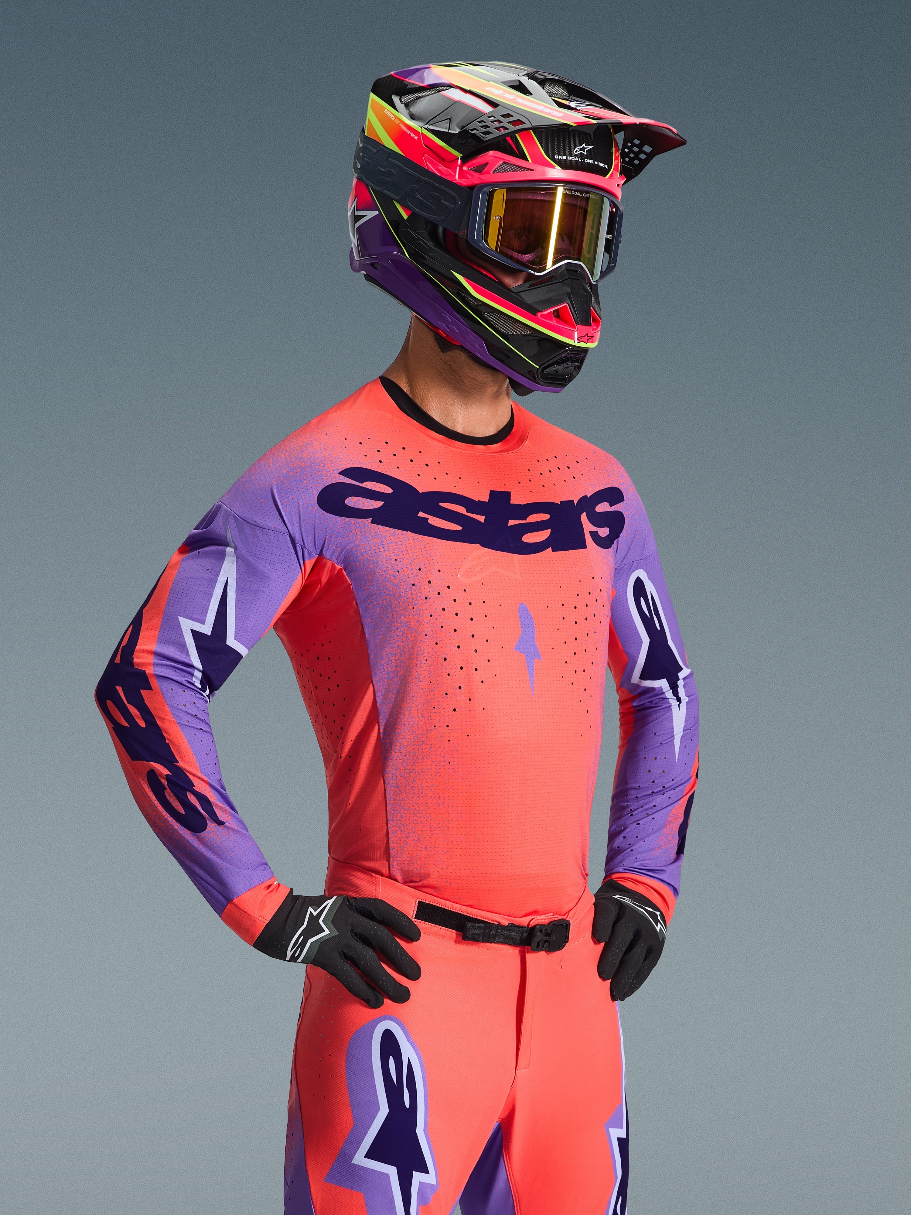 Pilota di motocross con maglia Alpinestars Supertech Scenz 2026, color corallo e viola con logo 'astars' sul petto, abbigliamento coordinato con pantaloni corallo e casco viola e corallo, con perforazioni laser per il fuoristrada.