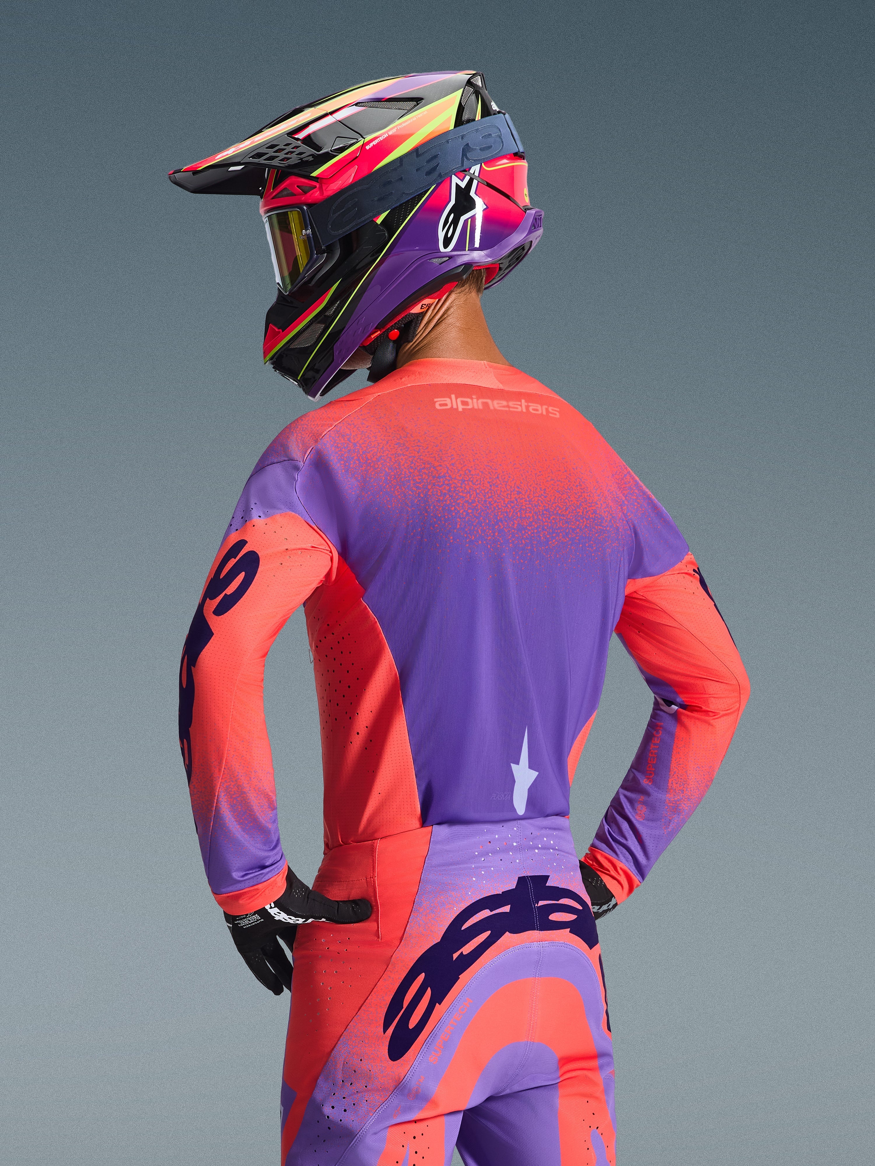 Pilota di motocross visto da dietro con la maglia Alpinestars Supertech Scenz 2026 in Hot Coral e Purple, caratterizzata da un vivace design sfumato e puntinato, abbinata a pantaloni da motocross coordinati e un casco da corsa colorato, abbigliamento professionale per prestazioni fuoristrada.