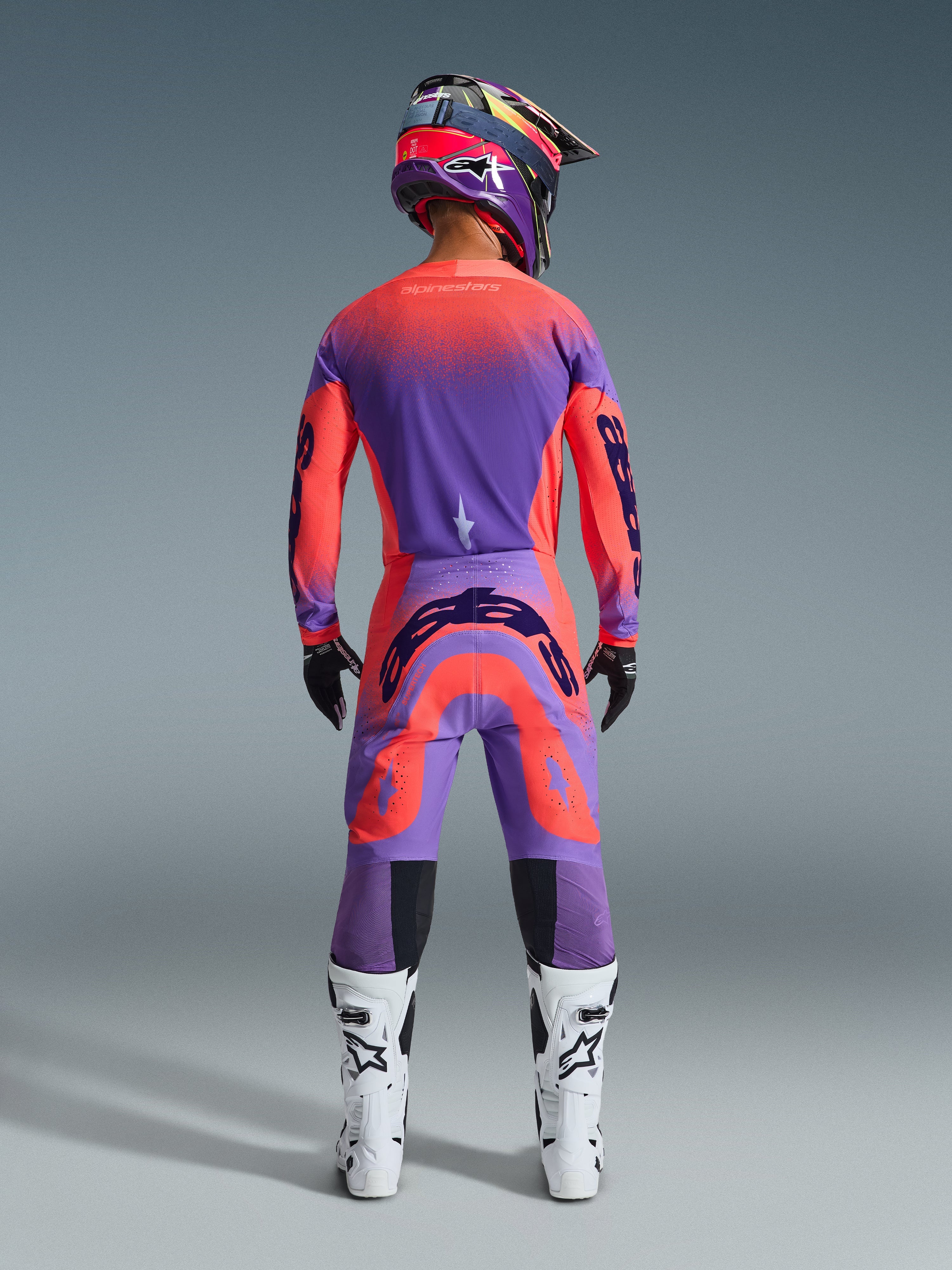 Pilota di motocross visto da dietro con il completo Supertech Scenz 2026 in Hot Coral Purple, maglia e pantaloni dai colori vivaci con transizione sfumata, loghi Alpinestars, casco coordinato, stivali bianchi e neri e guanti neri, abbigliamento professionale per gare off-road.