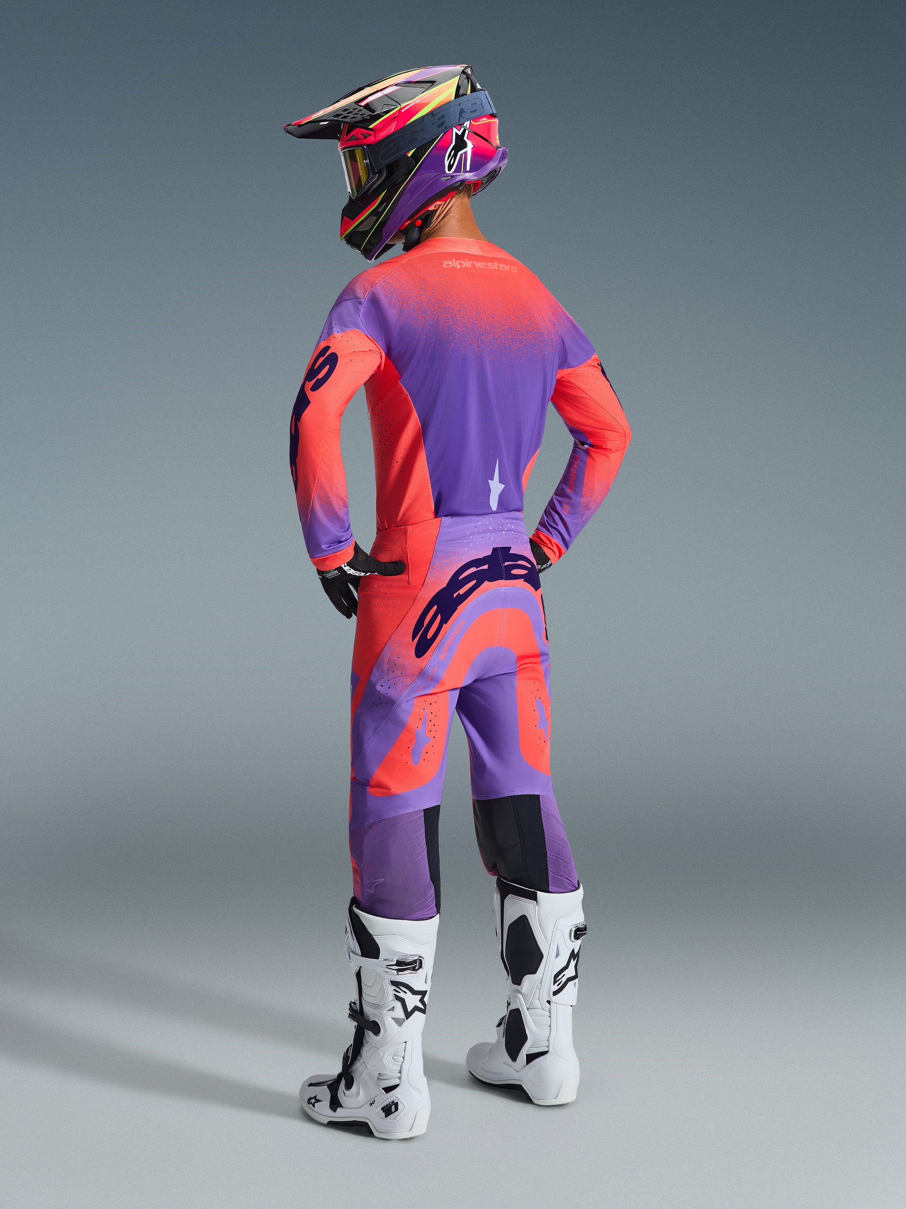 Pilota di motocross visto di spalle con il completo Alpinestars Supertech Scenz 2026, maglia e pantaloni viola e rosa corallo, grande logo Alpinestars sulla seduta e sulle maniche, casco coordinato viola e corallo, stivali bianchi e neri, abbigliamento professionale da gara coordinato