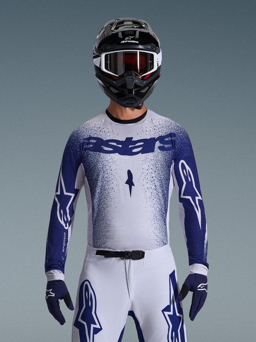 Pilota di motocross con maglia Alpinestars Supertech Scenz 2026, grigio chiaro e blu scuro con design a sfumatura puntinata e logo 'astars' in evidenza. Indossata con pantaloni da motocross bianchi coordinati, guanti blu scuro e casco nero con maschera, completo per gare professionistiche di fuoristrada.