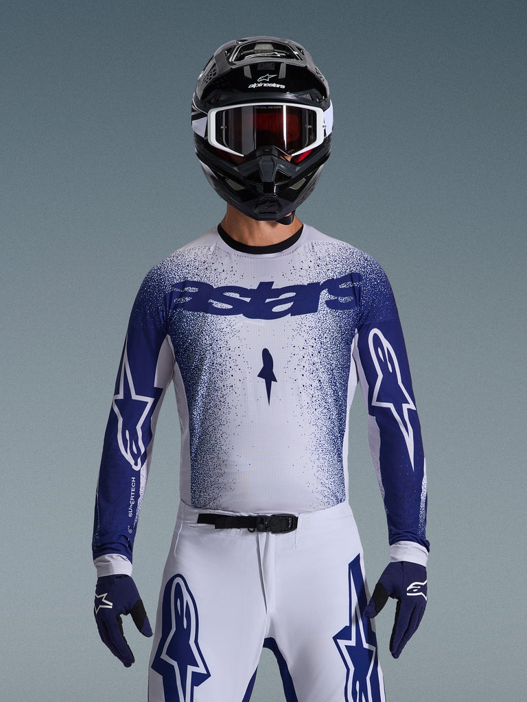 Pilota di motocross con maglia Alpinestars Supertech Scenz 2026, grigio chiaro e blu scuro con design a sfumatura puntinata e logo 'astars' in evidenza. Indossata con pantaloni da motocross bianchi coordinati, guanti blu scuro e casco nero con maschera, completo per gare professionistiche di fuoristrada.
