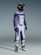 Pilota di motocross con abbigliamento completo da gara professionale, maglia Supertech Scenz 2026 in grigio chiaro e blu scuro, pantaloni da motocross coordinati con grafiche a stella, casco nero con maschera rossa, stivali tecnici bianchi e neri, guanti blu e bianchi coordinati per gare fuoristrada