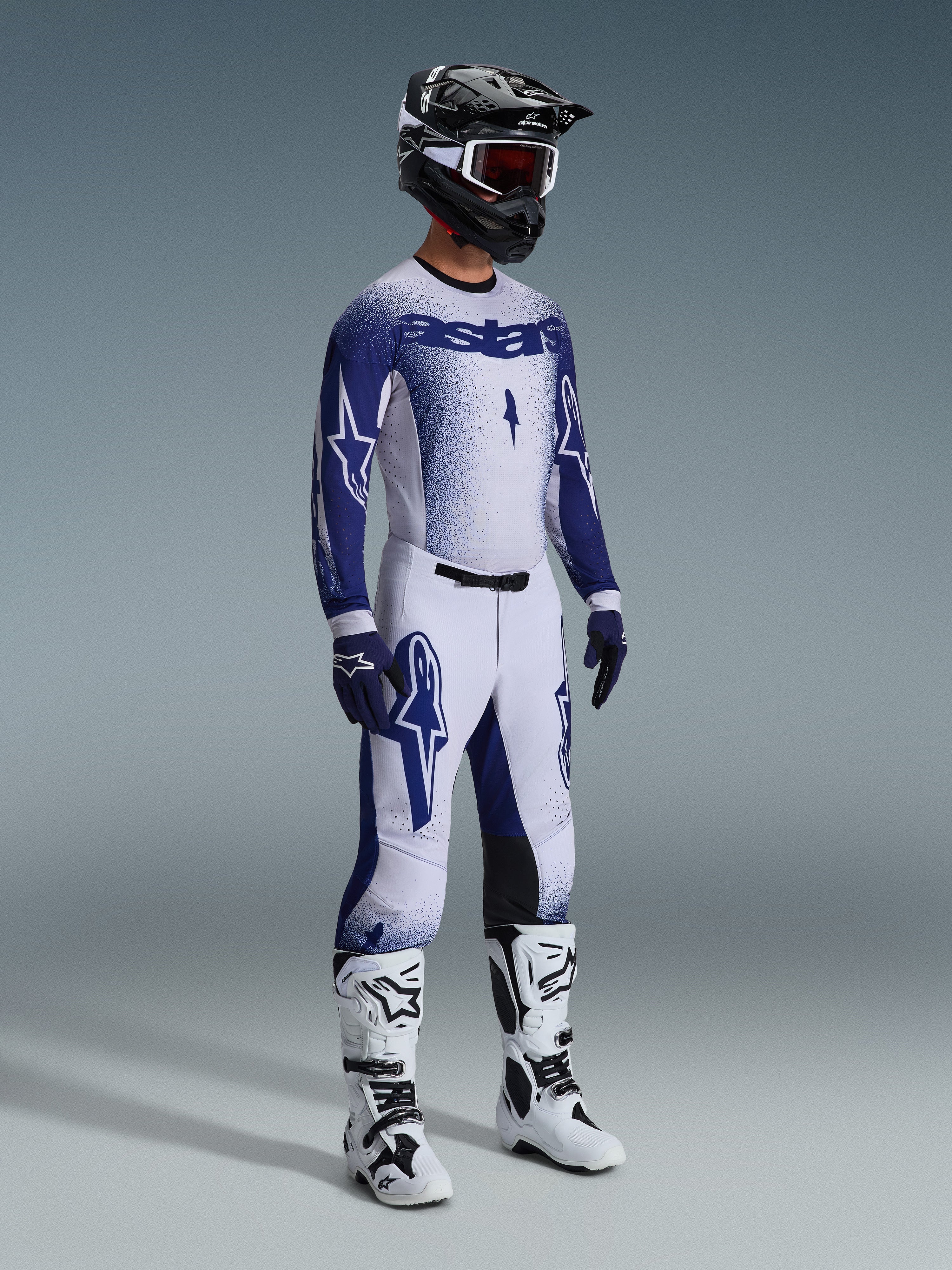 Pilota di motocross con abbigliamento completo da gara professionale, maglia Supertech Scenz 2026 in grigio chiaro e blu scuro, pantaloni da motocross coordinati con grafiche a stella, casco nero con maschera rossa, stivali tecnici bianchi e neri, guanti blu e bianchi coordinati per gare fuoristrada