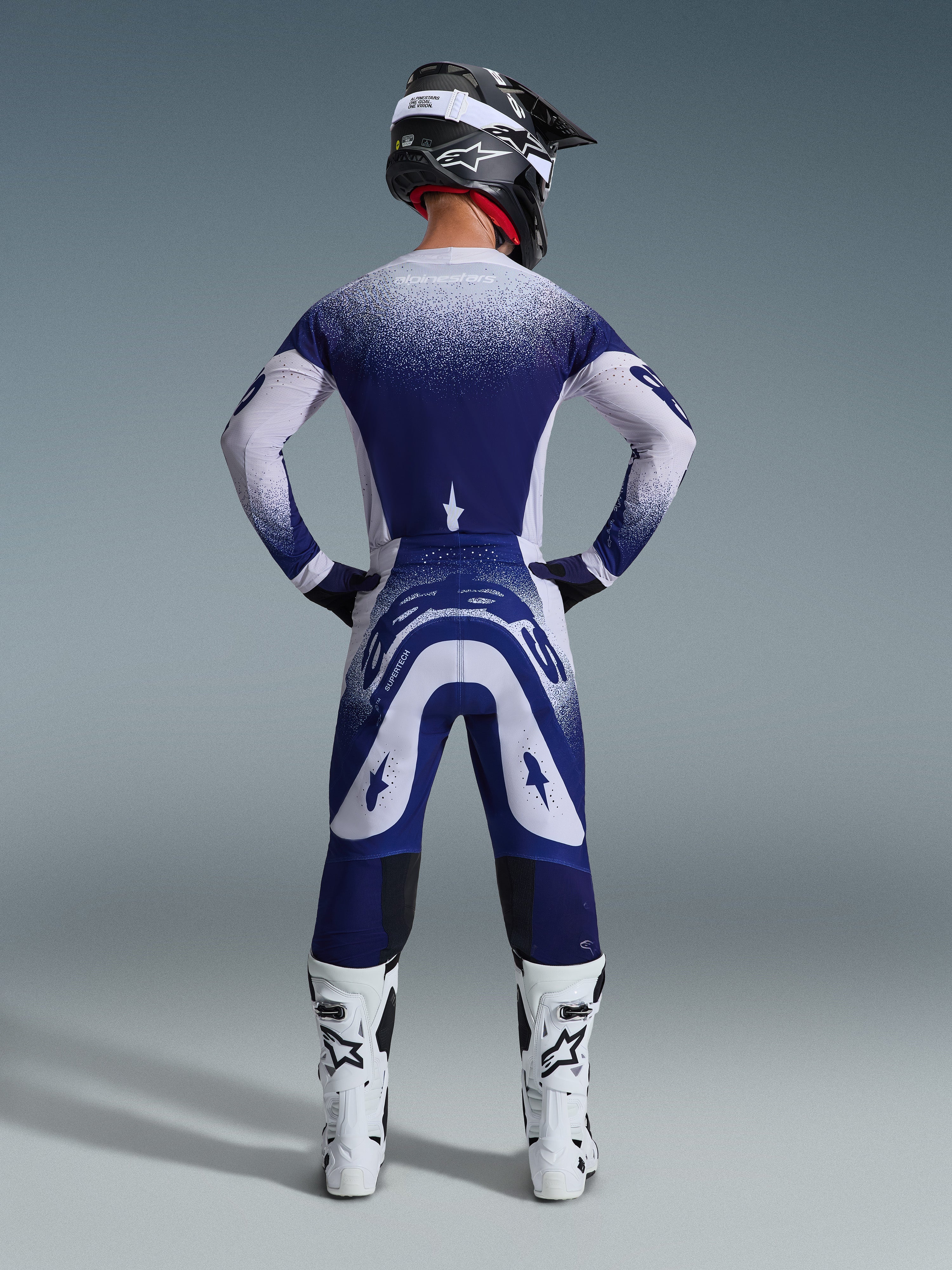 Alpinestars 2026 Supertech Scenz Gear Set, vista posteriore di un pilota di motocross con maglia e pantaloni da corsa blu e bianchi, effetto gradiente puntinato, indossato con stivali da motocross bianchi e casco nero, abbigliamento professionale da motocross
