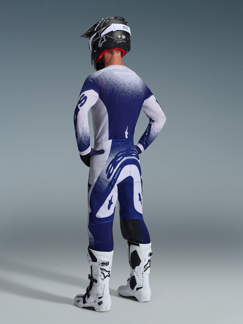 Vista posteriore di un pilota di motocross che indossa la maglia Alpinestars Supertech Scenz 2026 in Grigio Chiaro e Blu Scuro, con design a gradiente punteggiato, coordinata con pantaloni abbinati e stivali da motocross bianchi, abbigliamento tecnico professionale da gara.