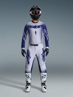 Pilota di motocross con abbigliamento da gara completo, maglia Supertech Scenz 2026 in grigio chiaro e blu scuro, pantaloni grigio chiaro coordinati con loghi blu, casco nero e bianco con maschera rossa, stivali bianchi e neri, guanti blu scuro, outfit coordinato per gare fuoristrada