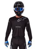 Alpinestars 2025 Racer Graphite Jersey, abbigliamento da motocross, nera con loghi grigi, indossata con casco nero, occhiali blu e guanti blu e grigi, design performante con loghi su petto e braccia