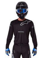 Alpinestars 2025 Racer Graphite Jersey, abbigliamento da motocross, nera con loghi grigi, indossata con casco nero, occhiali blu e guanti blu e grigi, design performante con loghi su petto e braccia